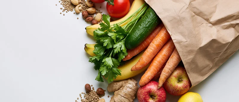 Brown paper grocery bag tipped over spilling fresh vegetables and fruits on white surface