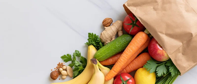 Brown paper grocery bag tipped over spilling fresh vegetables and fruits on white marble surface