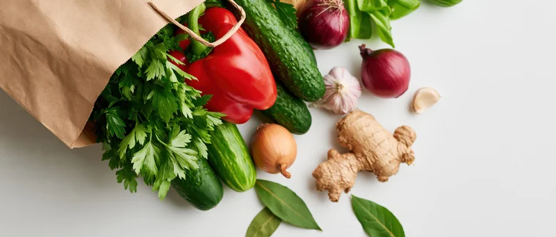 Brown paper bag spilling fresh vegetables and herbs on white surface