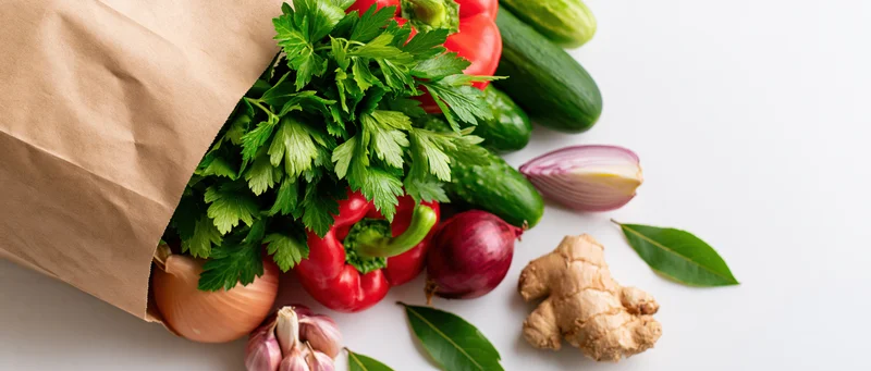 Brown paper grocery bag tipped over spilling fresh vegetables and herbs on white surface