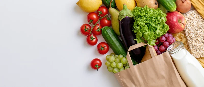 Brown paper grocery bag tipped on its side with fresh vegetables and fruits spilling out onto white surface