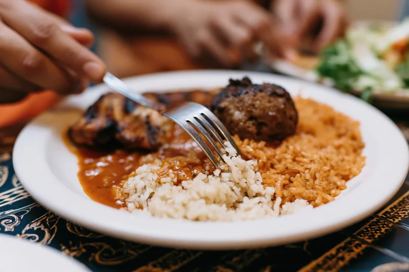 Fork pressing into a plate of grilled chicken and rice with sauce at a dining table