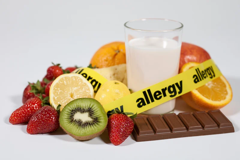 Glass of milk and assorted fresh fruits with chocolate bar surrounded by yellow allergy caution tape on white background
