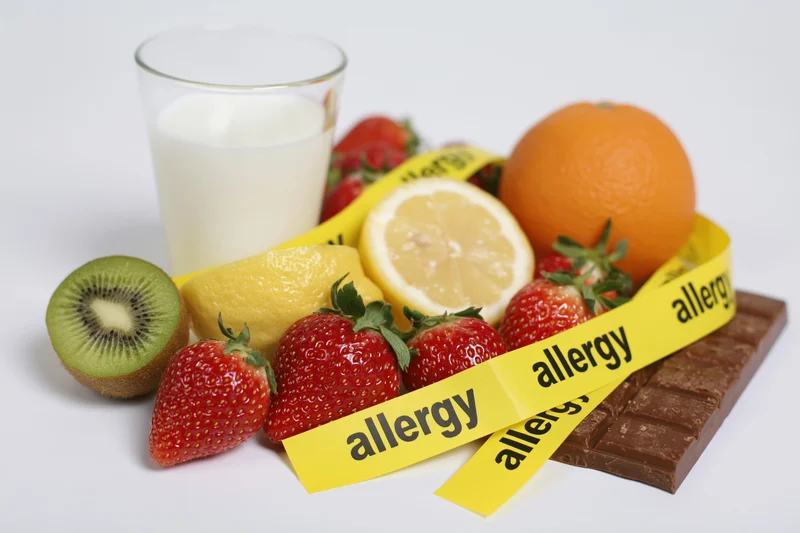 Various fresh fruits, milk, and chocolate arranged with yellow allergy warning tape on white background
