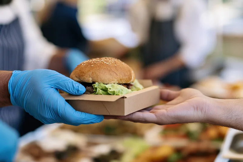 Gloved hand passing a burger in a paper box to another hand at an outdoor food event