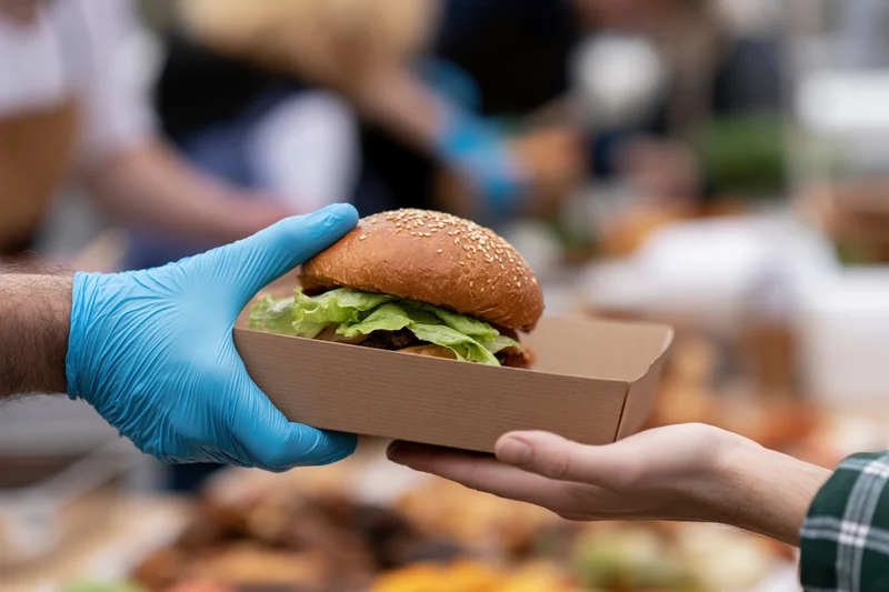 Gloved hand passing a burger in a paper box to a customer at an outdoor food event