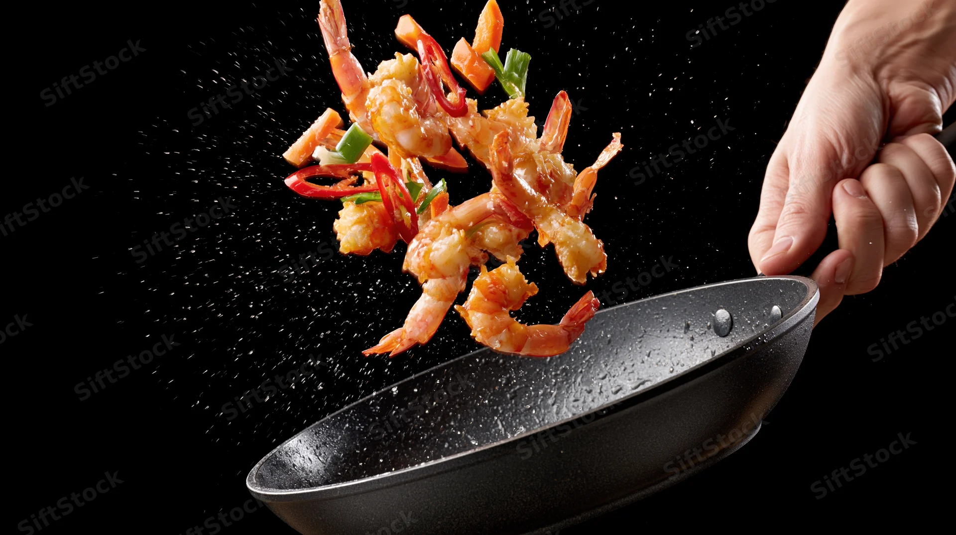 Hand tossing shrimp and red chili slices in a skillet against black background