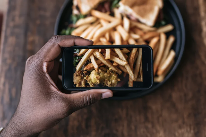 Hand holding smartphone capturing photo of fries and sandwich on black plate over wooden table