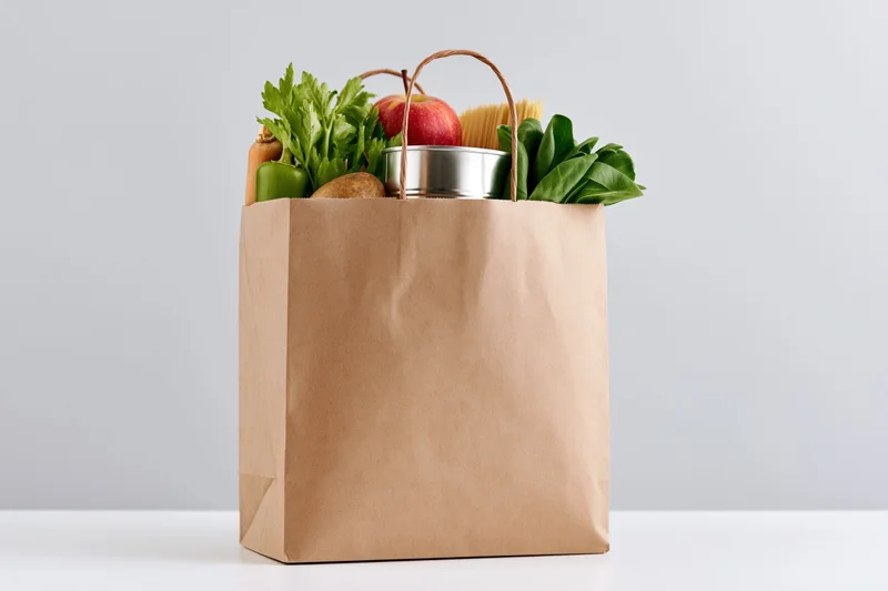 Kraft paper grocery bag filled with fresh vegetables and fruit on white surface