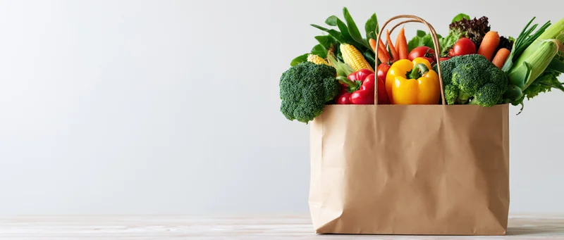 Kraft paper bag overflowing with fresh vegetables on a white surface