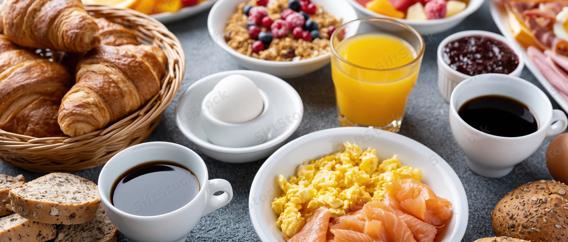 Continental breakfast spread on a table with croissants, coffee, eggs, salmon, cereal, fruit, and toast