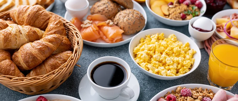 Continental breakfast spread on a table with croissants, coffee, scrambled eggs, orange juice, and assorted pastries and fruits