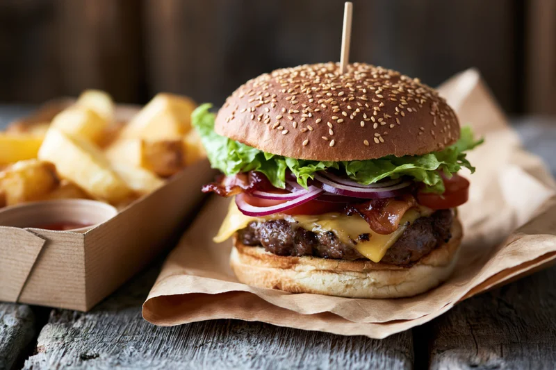 Loaded cheeseburger with lettuce tomato red onion bacon and melted cheese on sesame bun served with fries on wooden table
