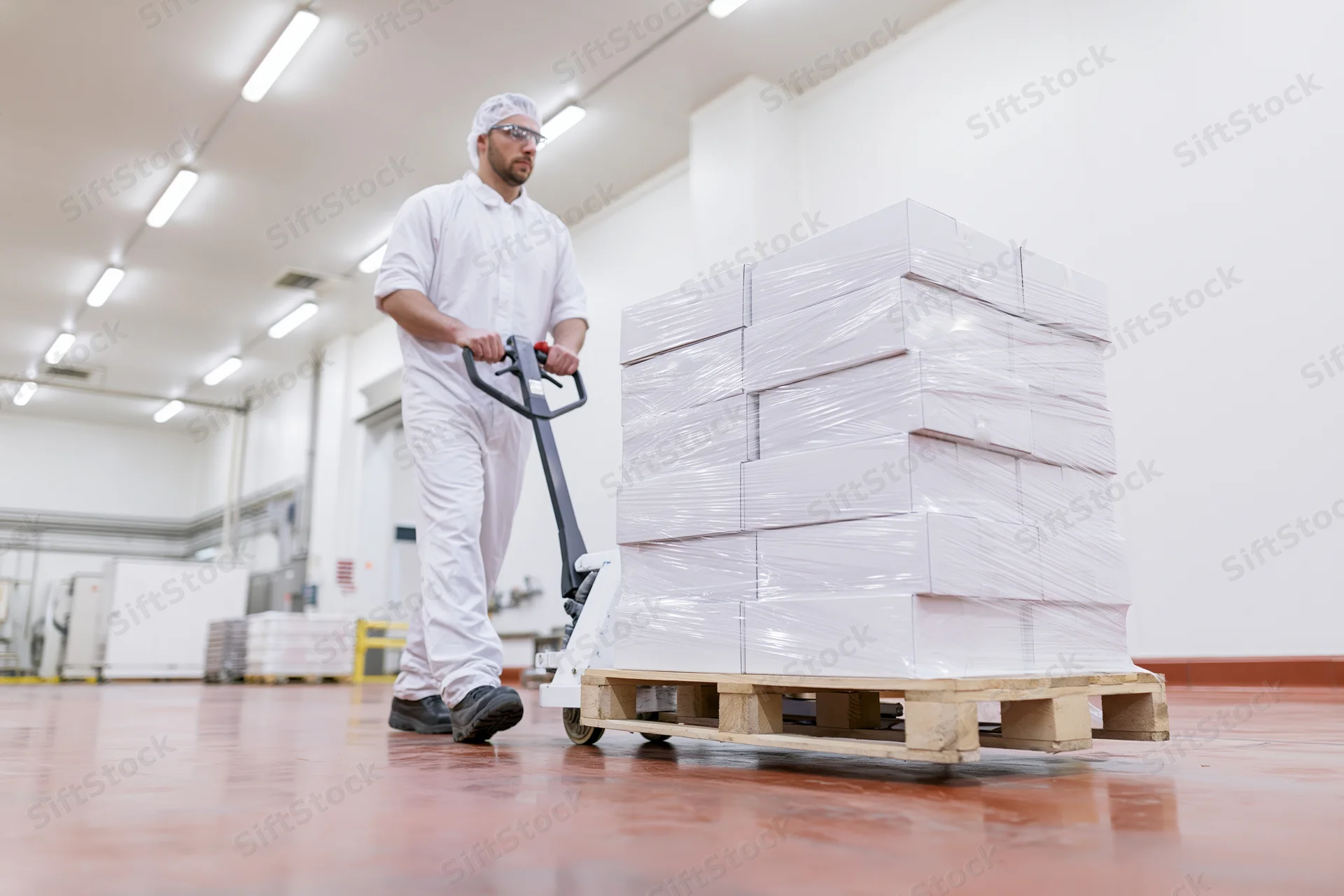 Male worker in white hygiene suit pushing pallet jack with stacked boxes in food factory warehouse