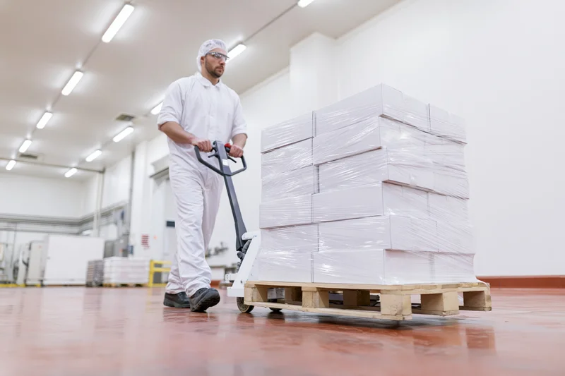 Male worker in white hygiene suit pushing pallet jack with stacked boxes in food factory warehouse