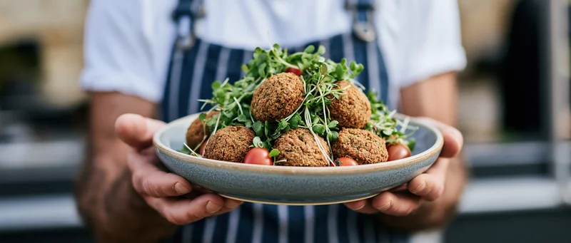 Man in denim apron holding ceramic plate with meatballs and microgreens