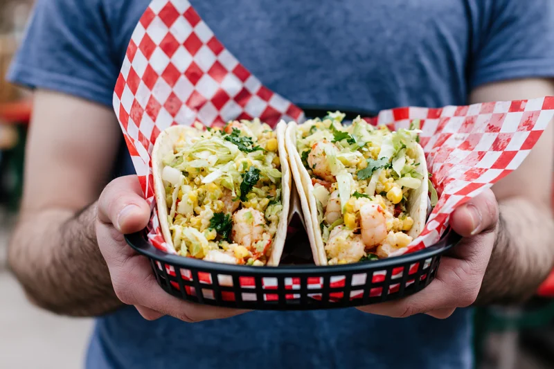 Man holding two loaded shrimp tacos in a basket lined with red and white checkered paper