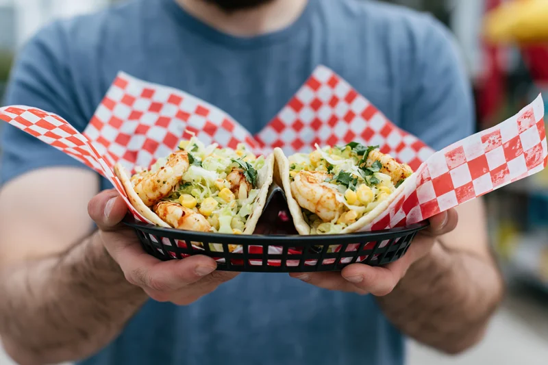 Man holding two shrimp tacos in a basket lined with checkered paper