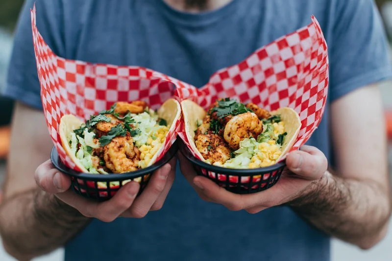 Man holding two shrimp tacos in baskets lined with red and white checkered paper