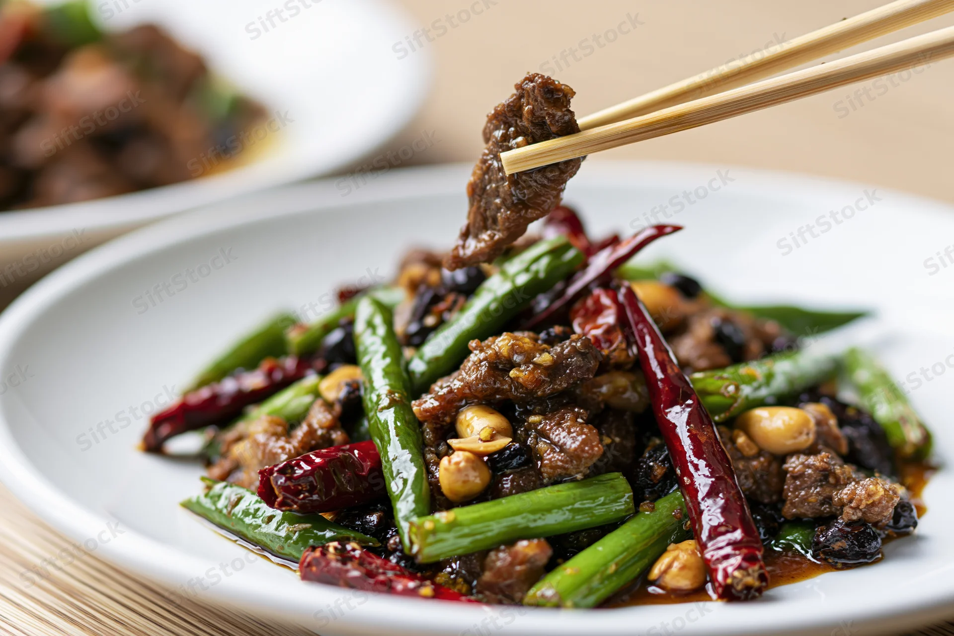 Chopsticks lifting a piece of stir-fried beef above a plate of vegetables and sauce