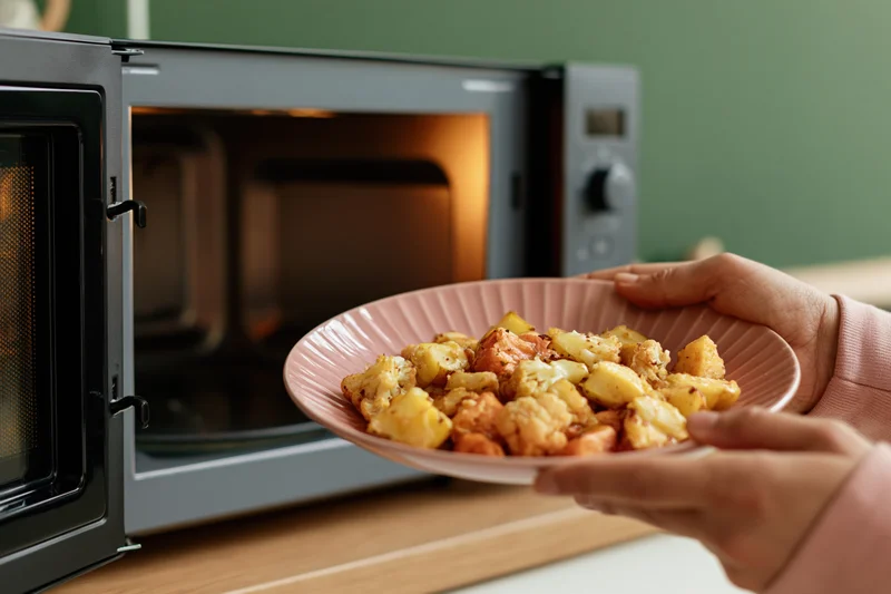 Hands holding a pink plate of roasted vegetables and potatoes in front of an open microwave on a kitchen countertop