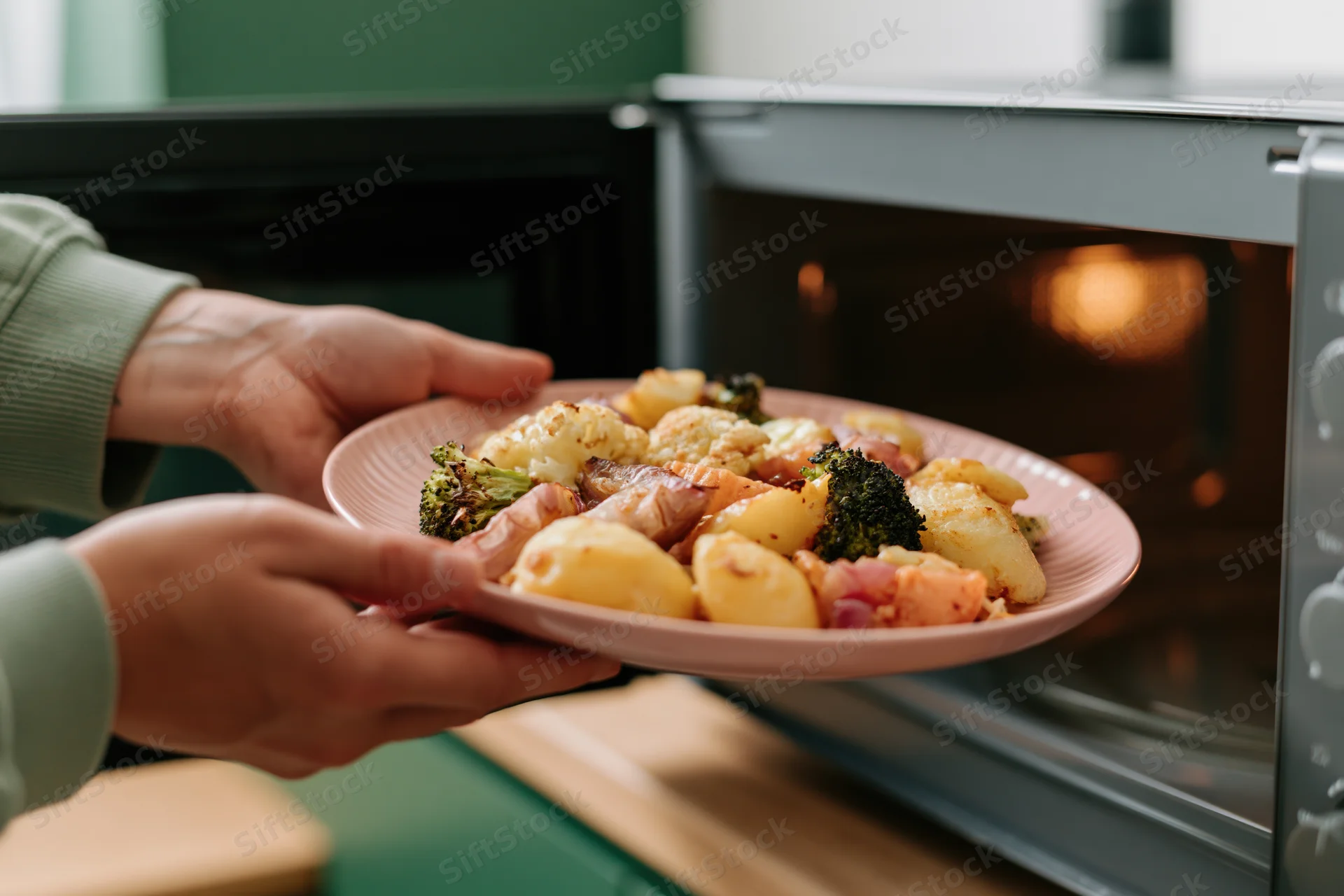 Hands holding a pink plate of roasted vegetables and potatoes near an open microwave in a kitchen
