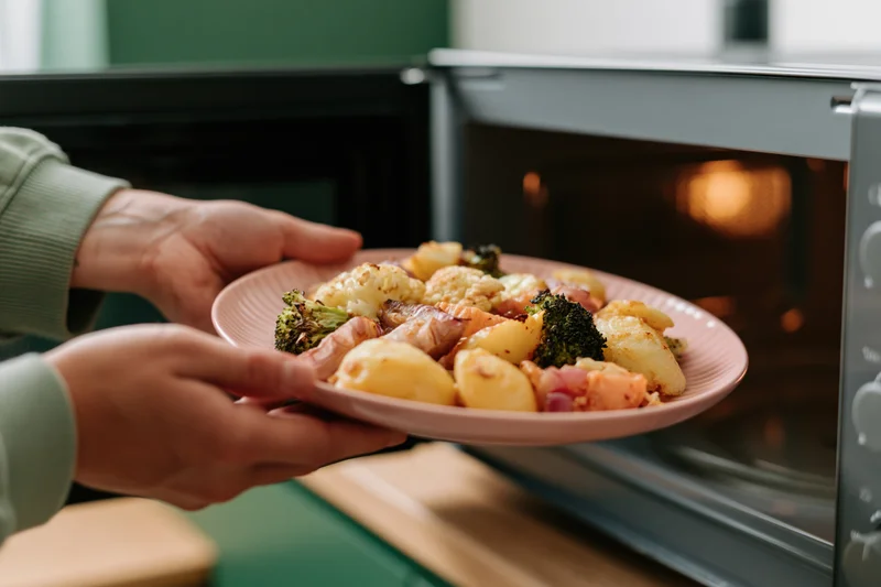 Hands holding a pink plate of roasted vegetables and potatoes near an open microwave in a kitchen