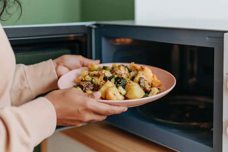 Woman placing a pink plate of roasted vegetables and potatoes into a microwave in a kitchen