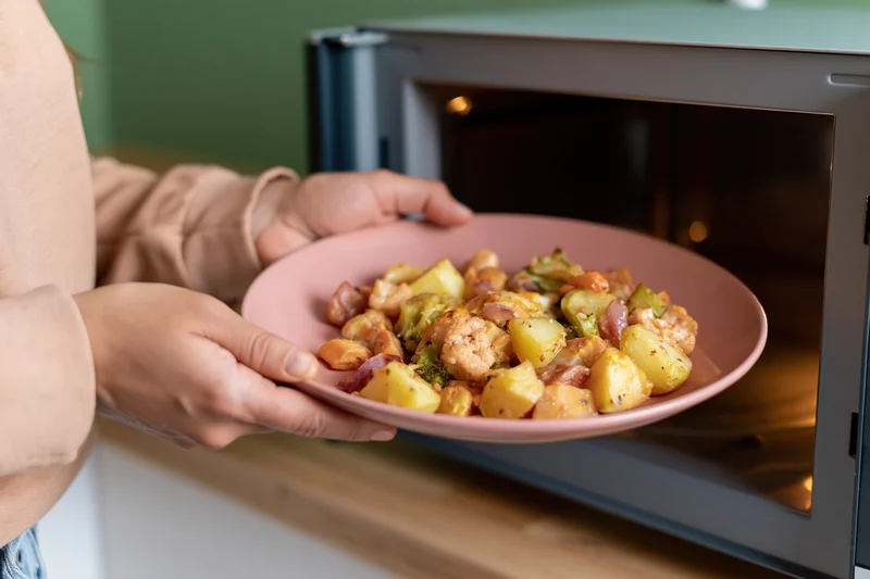 Woman placing a pink plate of roasted vegetables and potatoes into a microwave on a kitchen countertop
