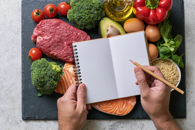Hands holding open blank spiral notebook over arranged fresh food ingredients on slate board