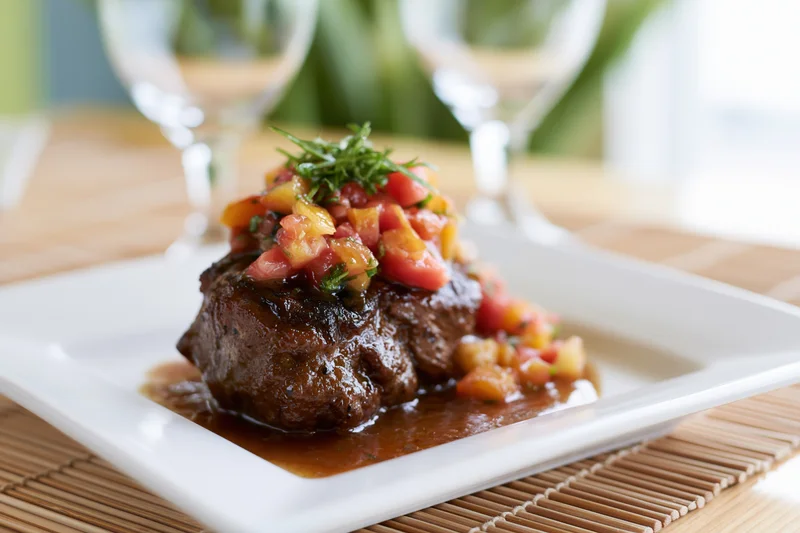 Seared beef steak topped with tomato herb sauce on a white square plate set on a bamboo placemat with wine glasses in the background