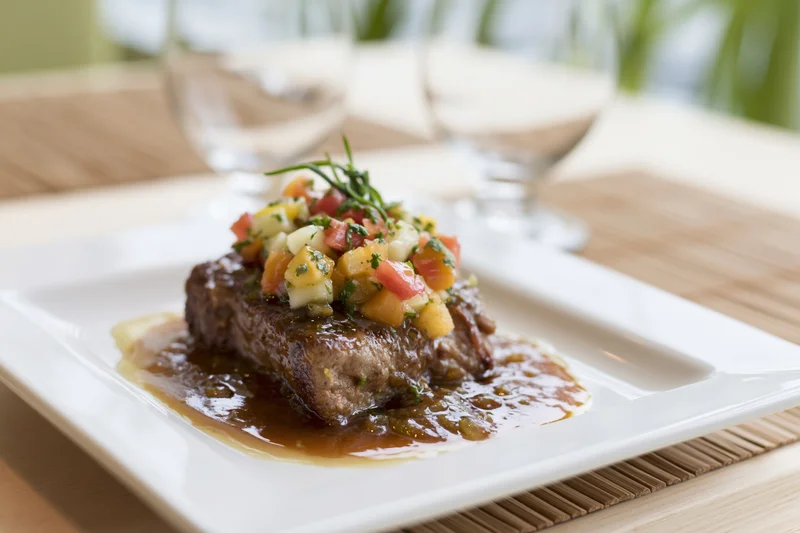 Seared steak with vegetable garnish and gravy on a white square plate at a restaurant table