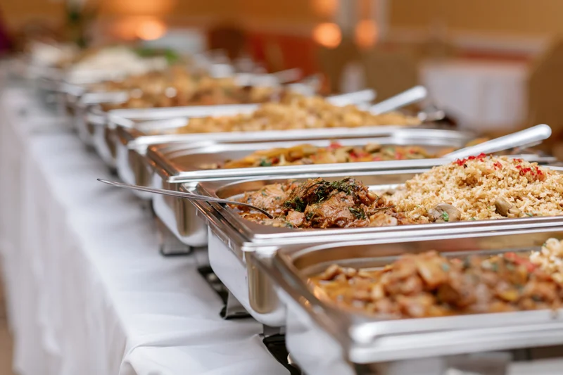 Row of stainless steel chafing dishes filled with hot food on a white tablecloth at a catered event