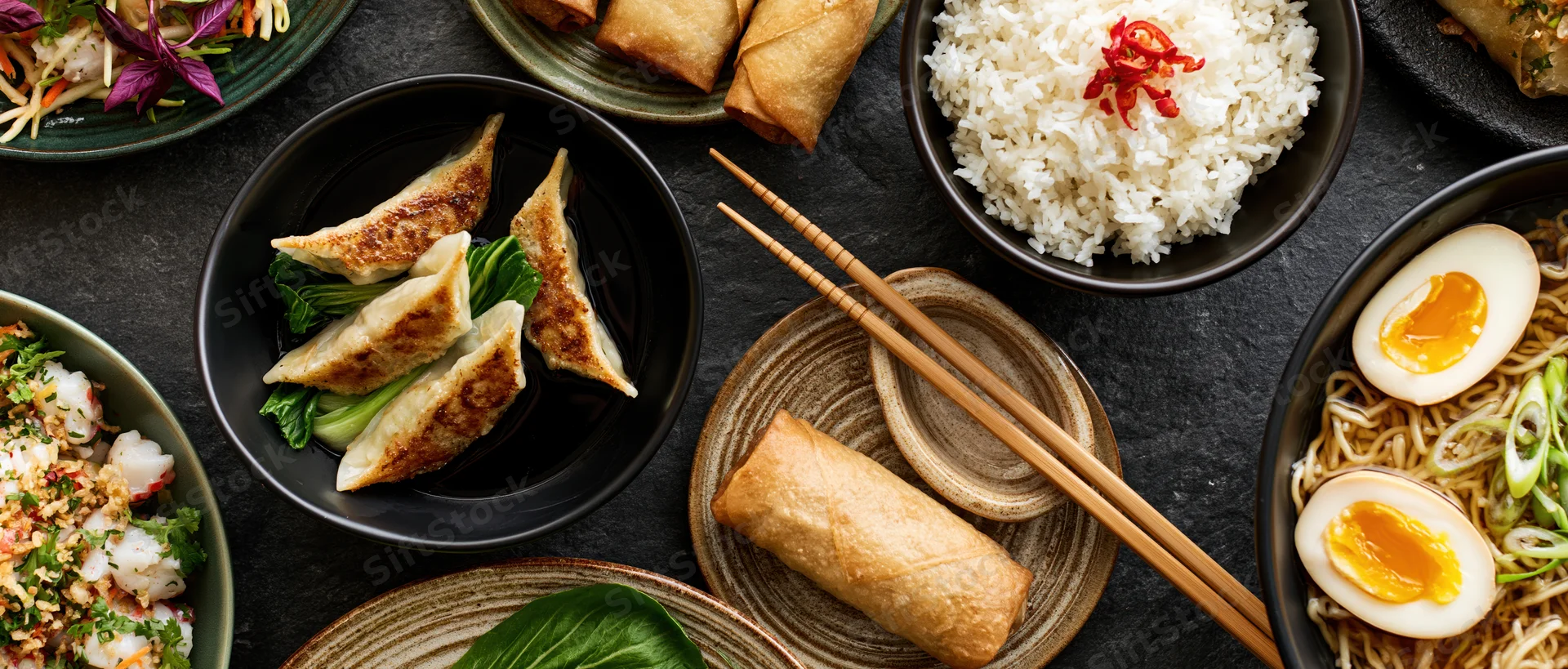 Spread of asian dishes including dumplings, spring rolls, rice, noodles, and boiled egg on dark stone surface with chopsticks and ceramic bowls