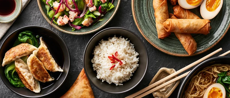 Asian dishes arranged on dark stone surface with bowls of rice, noodles, dumplings, spring rolls, salad, and chopsticks