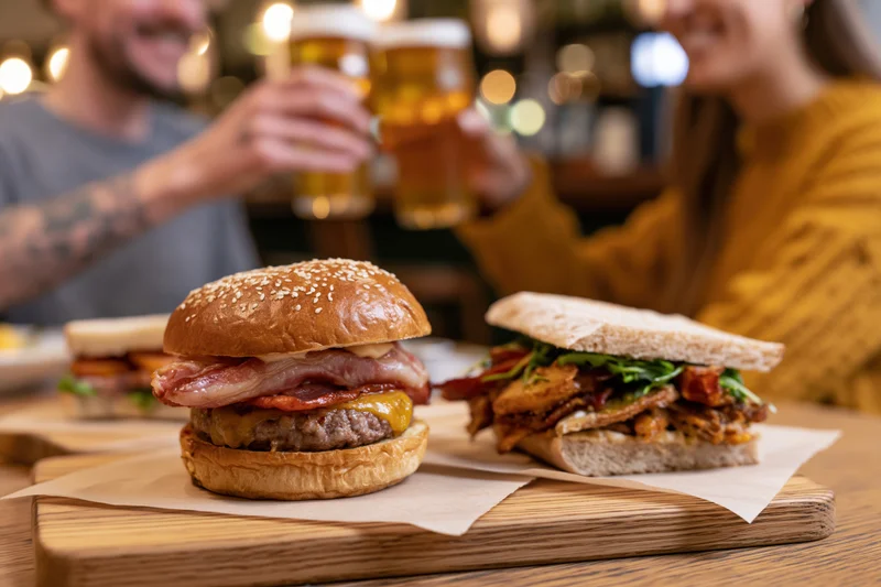 Burger and sandwich on wooden board with couple toasting beer in background