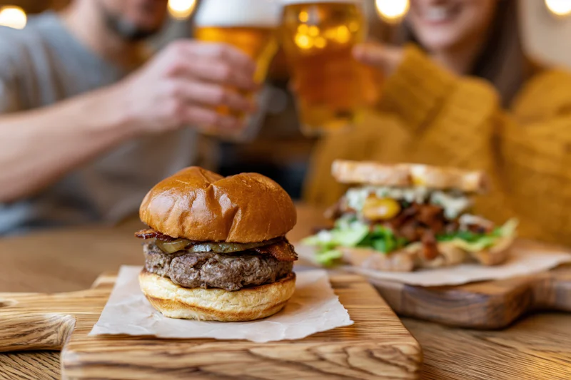 Stacked brioche burger with bacon and cheese on wooden board, couple toasting with beer in background