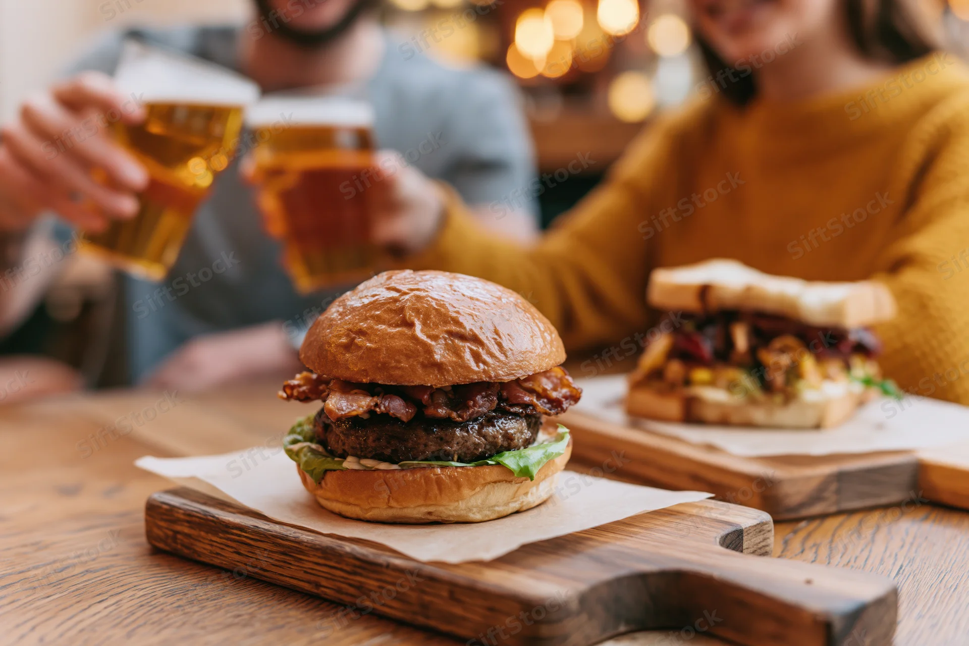 Stacked brioche burger with bacon and lettuce on wooden serving board, with couple toasting beer glasses in background