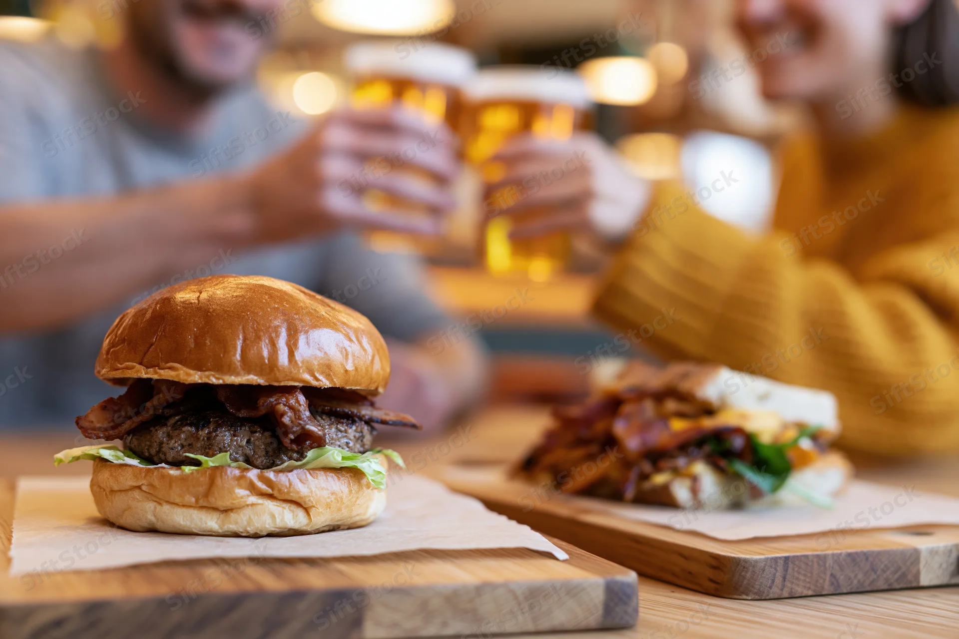 Stacked brioche burger with bacon and lettuce on wooden board in foreground, couple toasting with beer glasses in background at restaurant table