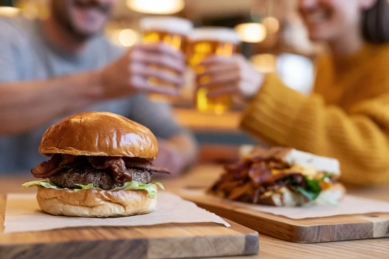 Stacked brioche burger with bacon and lettuce on wooden board in foreground, couple toasting with beer glasses in background at restaurant table