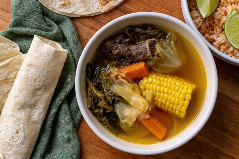 Bowl of mexican beef bone soup with corn, carrots, cabbage and tortillas on wooden table