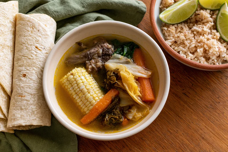 White ceramic bowl of mexican beef bone soup with vegetables and corn served with rice and tortillas on wooden table