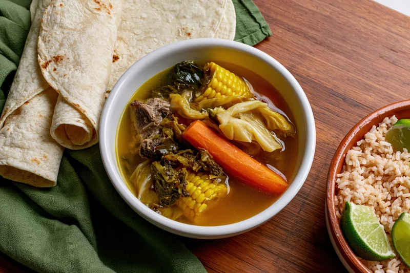 Bowl of mexican caldo de res soup with corn, carrot, cabbage, tortillas, rice, and lime on wooden table