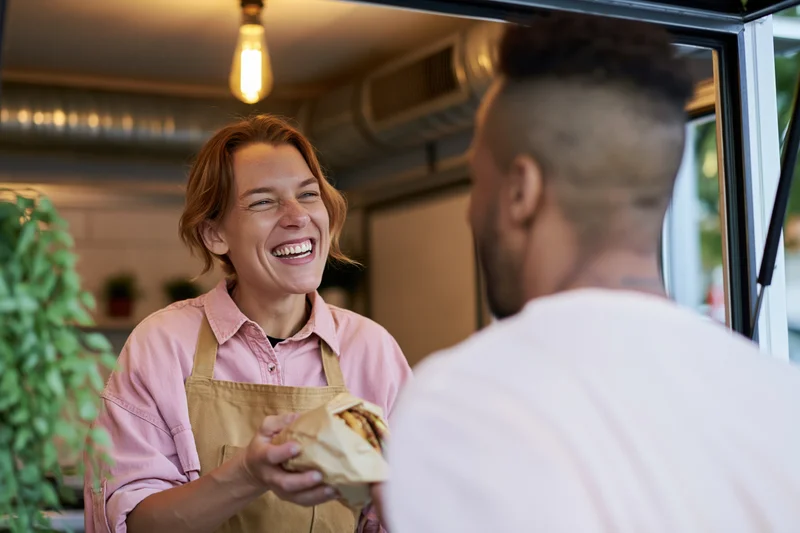 Woman in apron laughing while serving sandwich to customer at food truck window