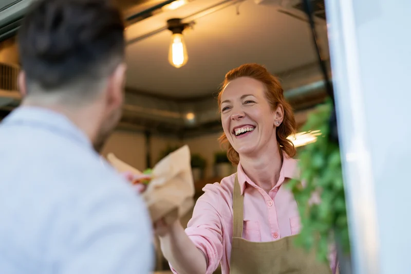 Woman in pink shirt and apron laughing while handing sandwich to customer inside food truck