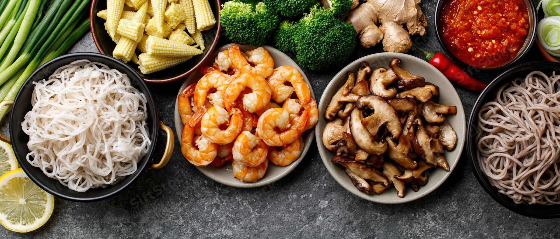 Shrimp, mushrooms, noodles, broccoli, corn, ginger, chili, lemon, green onions, and sauce arranged in bowls on a dark surface for Asian cooking