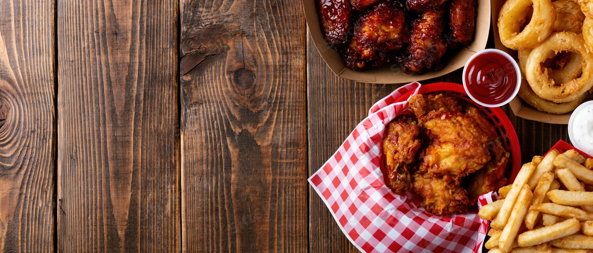 Assorted bar food including fried chicken, wings, onion rings and fries served in baskets on a wooden table with copyspace on the left