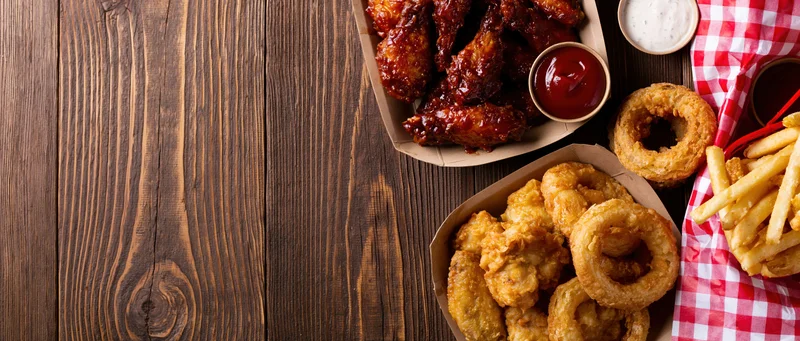 Assorted bar food including wings, onion rings and fries arranged on a wooden table with checkered napkin and sauces, copyspace on left