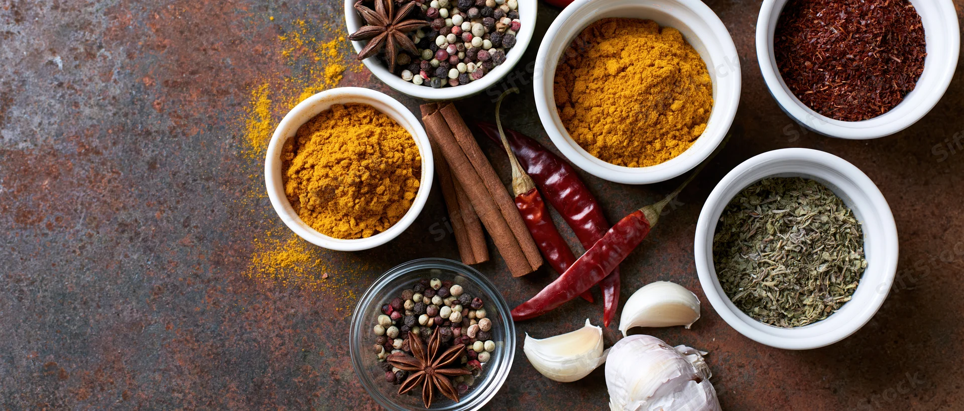 Assorted cooking spices in bowls and whole forms arranged on a dark rusted metal surface