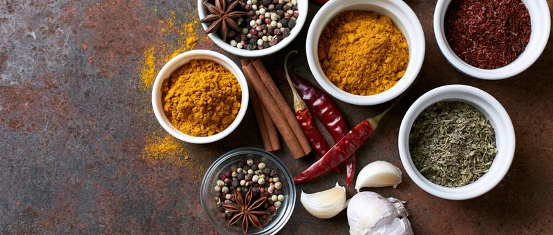 Assorted cooking spices in bowls and whole forms arranged on a dark rusted metal surface